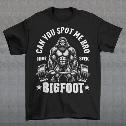 Black T-shirt featuring a muscular cartoon Bigfoot lifting a heavily loaded barbell. The graphic has bold white vintage-style lettering reading “CAN YOU SPOT ME BRO” above the Sasquatch, with “HIDE” and “SEEK” on either side and “BIGFOOT” beneath the weightlifter, flanked by stars. The design combines Bigfoot humor, gym culture, weightlifting, and cryptid themes.