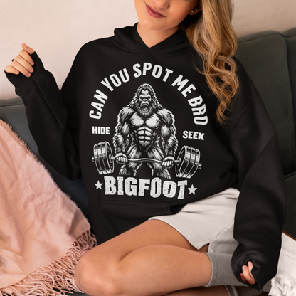 Black Hoodie featuring a muscular cartoon Bigfoot lifting a heavily loaded barbell. The graphic has bold white vintage-style lettering reading “CAN YOU SPOT ME BRO” above the Sasquatch, with “HIDE” and “SEEK” on either side and “BIGFOOT” beneath the weightlifter, flanked by stars. The design combines Bigfoot humor, gym culture, weightlifting, and cryptid themes.