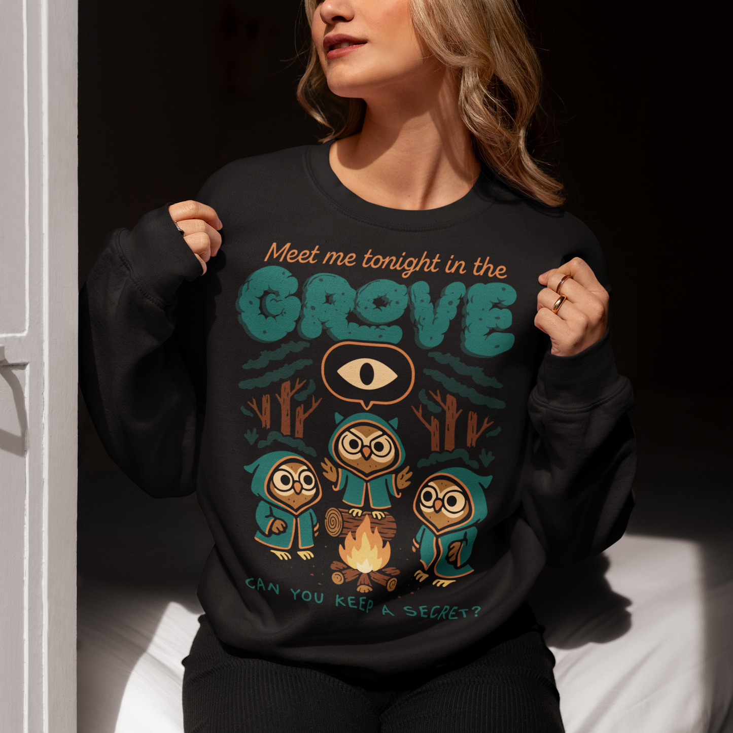 Black sweatshirt featuring three hooded owl characters gathered around a campfire in a dark forest. Above them is the text “Meet Me Tonight in the Grove” with an all-seeing eye symbol, and below reads “Can You Keep a Secret?” The design uses teal, orange, and cream tones in a spooky vintage cartoon style inspired by occult themes and secret societies.