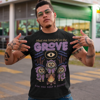 Black t-shirt featuring three hooded owl characters gathered around a campfire in a dark forest. Above them is the text “Meet Me Tonight in the Grove” with an all-seeing eye symbol, and below reads “Can You Keep a Secret?” The design uses teal, orange, and cream tones in a spooky vintage cartoon style inspired by occult themes and secret societies.