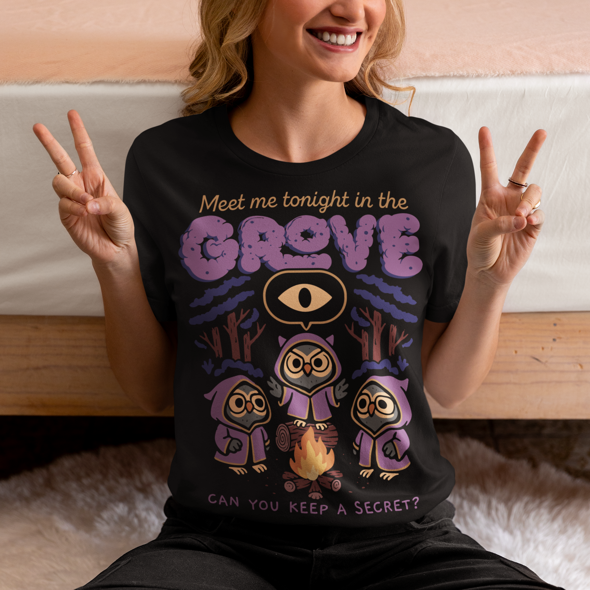 Black t-shirt featuring three hooded owl characters gathered around a campfire in a dark forest. Above them is the text “Meet Me Tonight in the Grove” with an all-seeing eye symbol, and below reads “Can You Keep a Secret?” The design uses teal, orange, and cream tones in a spooky vintage cartoon style inspired by occult themes and secret societies.