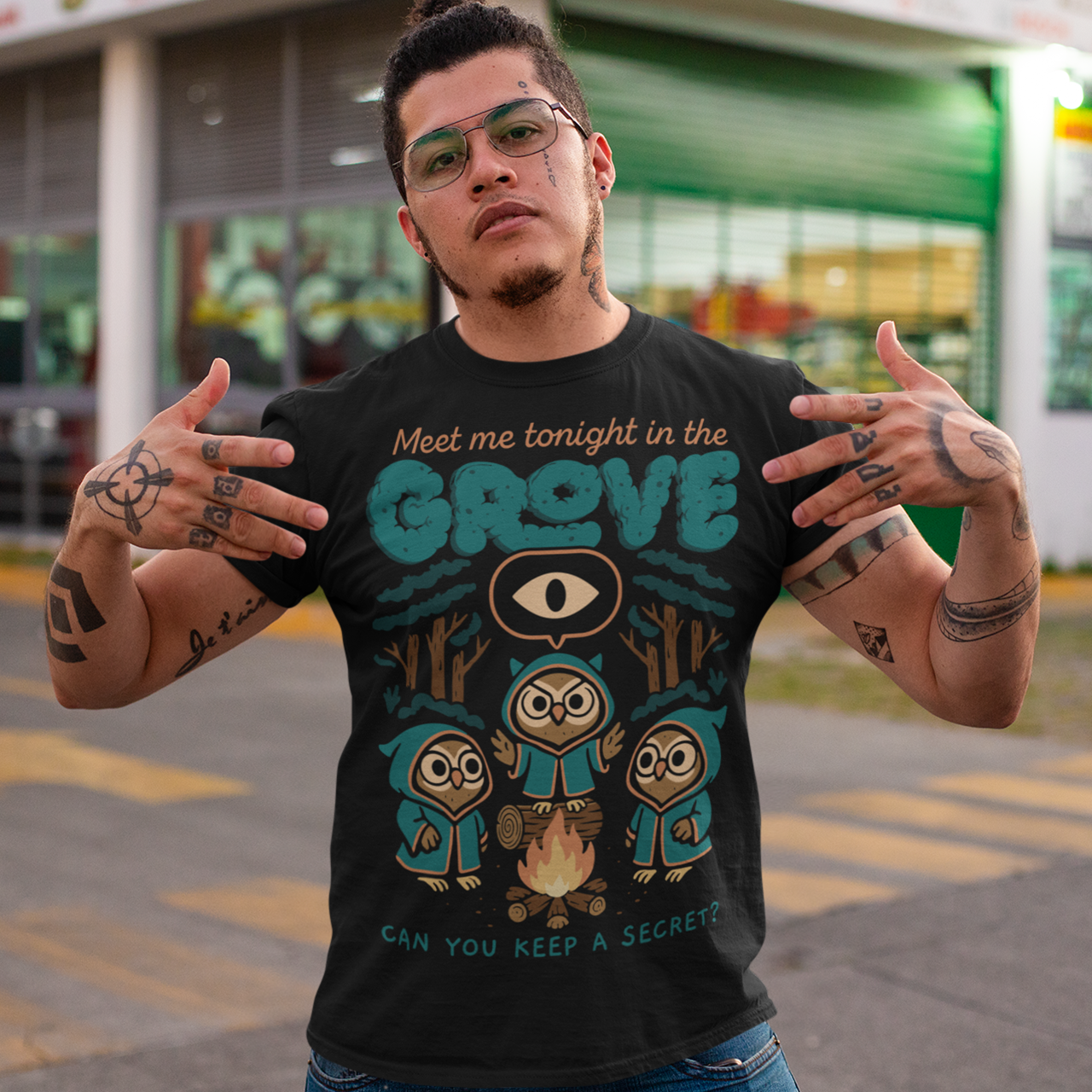 Black t-shirt featuring three hooded owl characters gathered around a campfire in a dark forest. Above them is the text “Meet Me Tonight in the Grove” with an all-seeing eye symbol, and below reads “Can You Keep a Secret?” The design uses teal, orange, and cream tones in a spooky vintage cartoon style inspired by occult themes and secret societies.