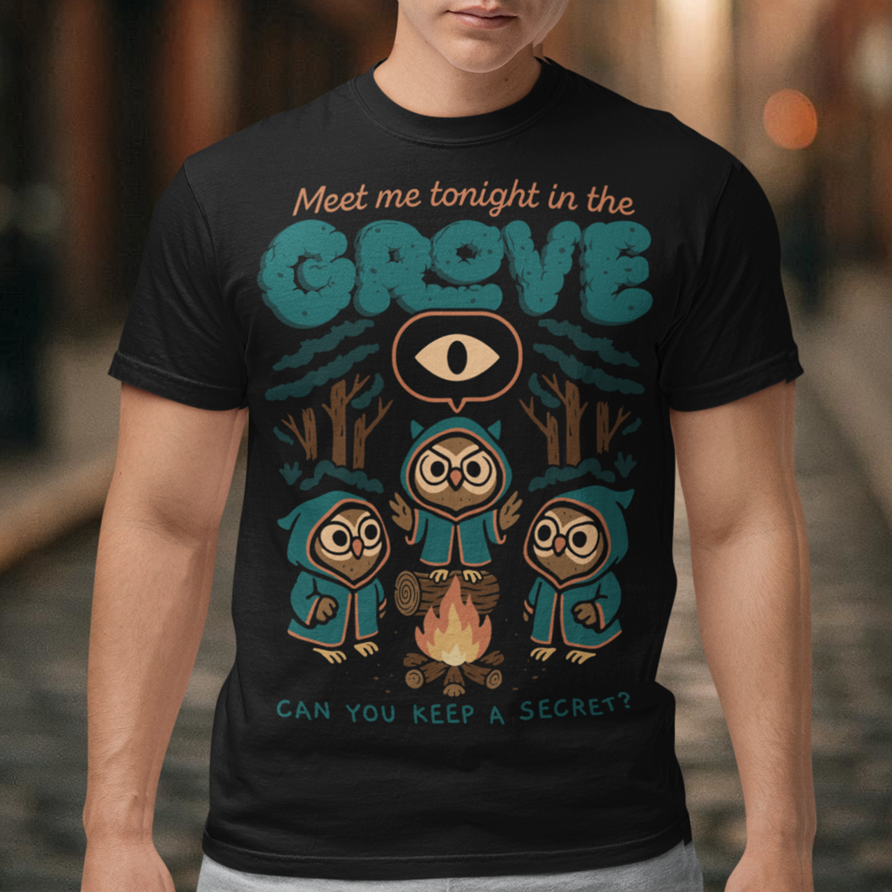 Black t-shirt featuring three hooded owl characters gathered around a campfire in a dark forest. Above them is the text “Meet Me Tonight in the Grove” with an all-seeing eye symbol, and below reads “Can You Keep a Secret?” The design uses teal, orange, and cream tones in a spooky vintage cartoon style inspired by occult themes and secret societies.