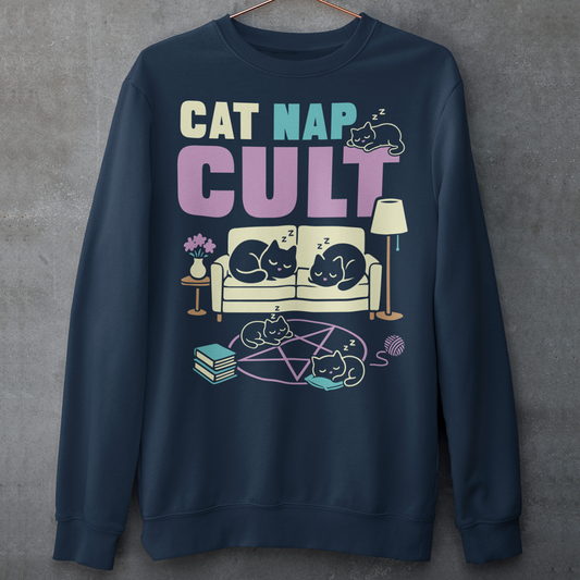 Navy sweatshirt with bold white text reading “Cat Nap Cult” above illustrations of sleeping cats lounging on a couch, books, and a pentagram-style ritual circle. The design includes cozy home details like a lamp, flowers, and yarn ball, creating a funny mix of occult humor and sleepy cat vibes in a minimalist monochrome style.