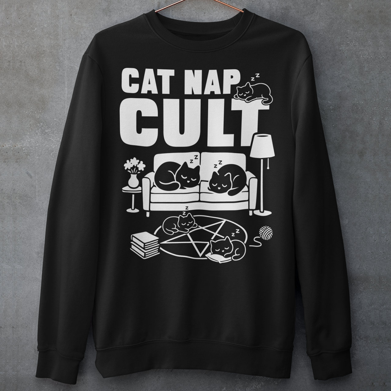 Black sweatshirt with bold white text reading “Cat Nap Cult” above illustrations of sleeping cats lounging on a couch, books, and a pentagram-style ritual circle. The design includes cozy home details like a lamp, flowers, and yarn ball, creating a funny mix of occult humor and sleepy cat vibes in a minimalist monochrome style.
