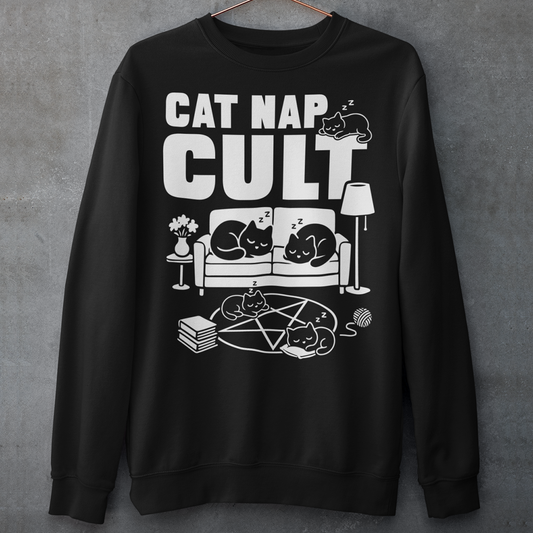 Black sweatshirt with bold white text reading “Cat Nap Cult” above illustrations of sleeping cats lounging on a couch, books, and a pentagram-style ritual circle. The design includes cozy home details like a lamp, flowers, and yarn ball, creating a funny mix of occult humor and sleepy cat vibes in a minimalist monochrome style.