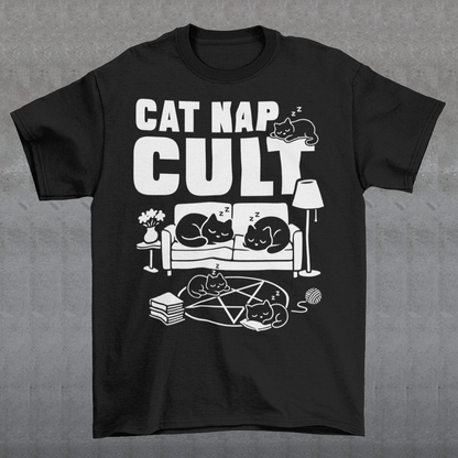 Black t-shirt with bold white text reading “Cat Nap Cult” above illustrations of sleeping cats lounging on a couch, books, and a pentagram-style ritual circle. The design includes cozy home details like a lamp, flowers, and yarn ball, creating a funny mix of occult humor and sleepy cat vibes in a minimalist monochrome style.
