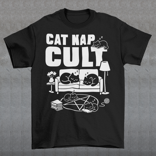 Black t-shirt with bold white text reading “Cat Nap Cult” above illustrations of sleeping cats lounging on a couch, books, and a pentagram-style ritual circle. The design includes cozy home details like a lamp, flowers, and yarn ball, creating a funny mix of occult humor and sleepy cat vibes in a minimalist monochrome style.