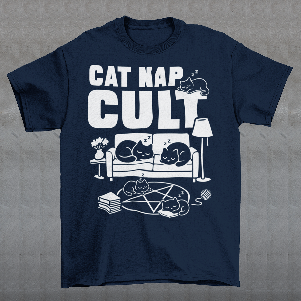 Navy t-shirt with bold white text reading “Cat Nap Cult” above illustrations of sleeping cats lounging on a couch, books, and a pentagram-style ritual circle. The design includes cozy home details like a lamp, flowers, and yarn ball, creating a funny mix of occult humor and sleepy cat vibes in a minimalist monochrome style.