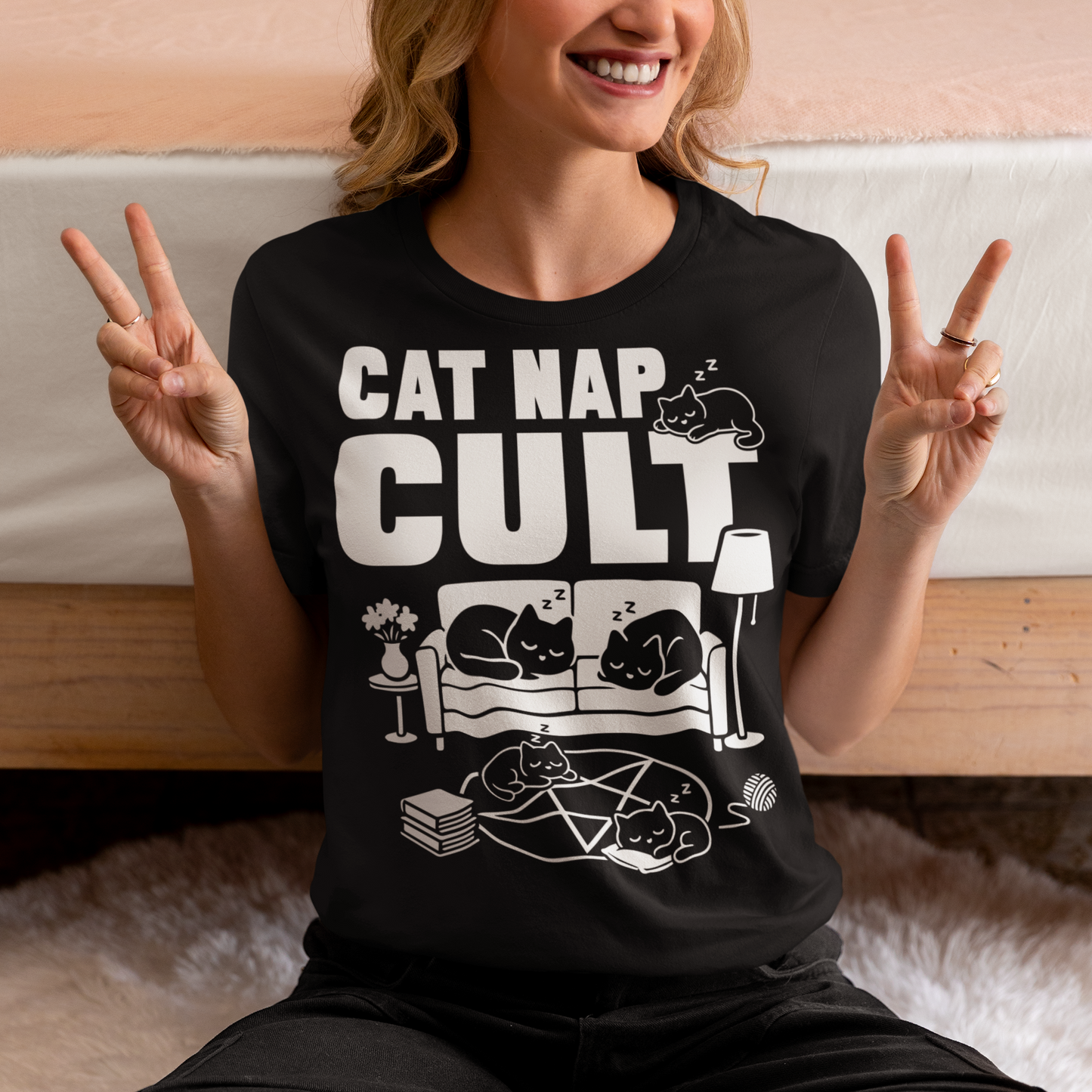 Black t-shirt with bold white text reading “Cat Nap Cult” above illustrations of sleeping cats lounging on a couch, books, and a pentagram-style ritual circle. The design includes cozy home details like a lamp, flowers, and yarn ball, creating a funny mix of occult humor and sleepy cat vibes in a minimalist monochrome style.