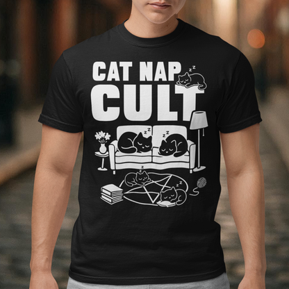 Black t-shirt with bold white text reading “Cat Nap Cult” above illustrations of sleeping cats lounging on a couch, books, and a pentagram-style ritual circle. The design includes cozy home details like a lamp, flowers, and yarn ball, creating a funny mix of occult humor and sleepy cat vibes in a minimalist monochrome style.
