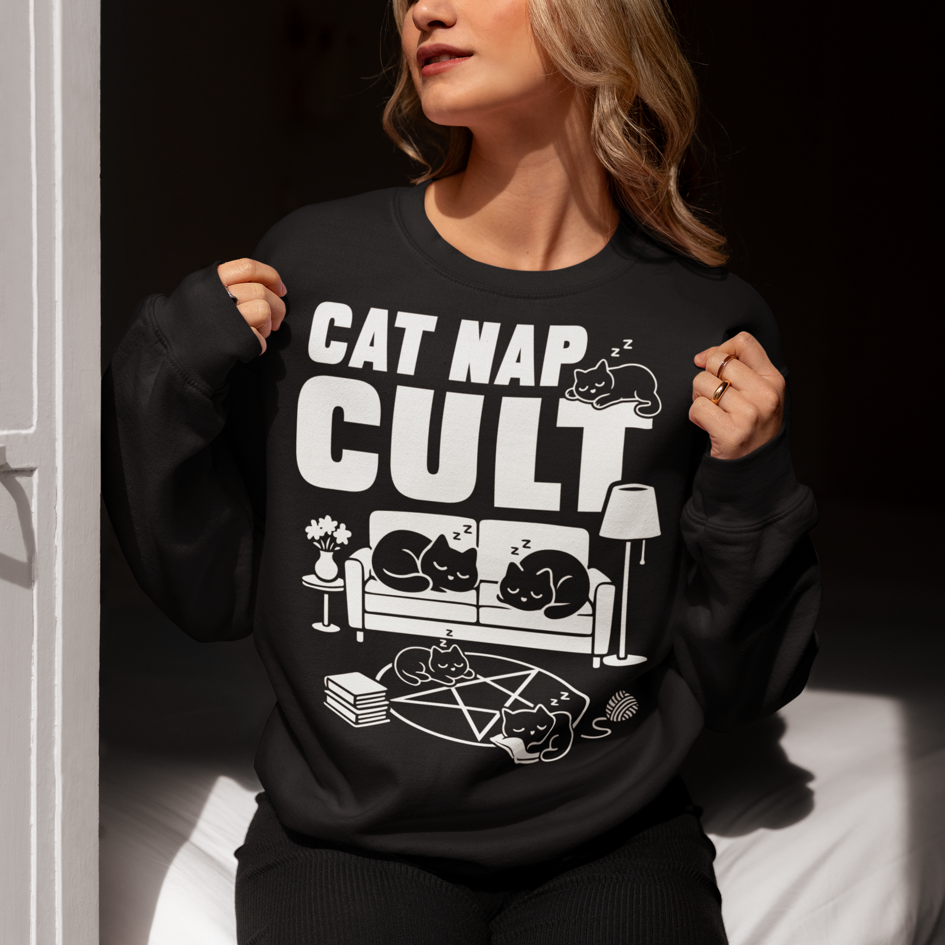 Black sweatshirt with bold white text reading “Cat Nap Cult” above illustrations of sleeping cats lounging on a couch, books, and a pentagram-style ritual circle. The design includes cozy home details like a lamp, flowers, and yarn ball, creating a funny mix of occult humor and sleepy cat vibes in a minimalist monochrome style.