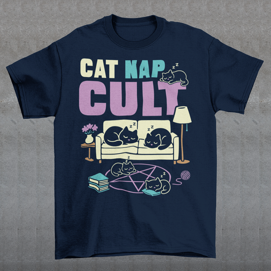 Navy t-shirt with bold white text reading “Cat Nap Cult” above illustrations of sleeping cats lounging on a couch, books, and a pentagram-style ritual circle. The design includes cozy home details like a lamp, flowers, and yarn ball, creating a funny mix of occult humor and sleepy cat vibes in a minimalist monochrome style.