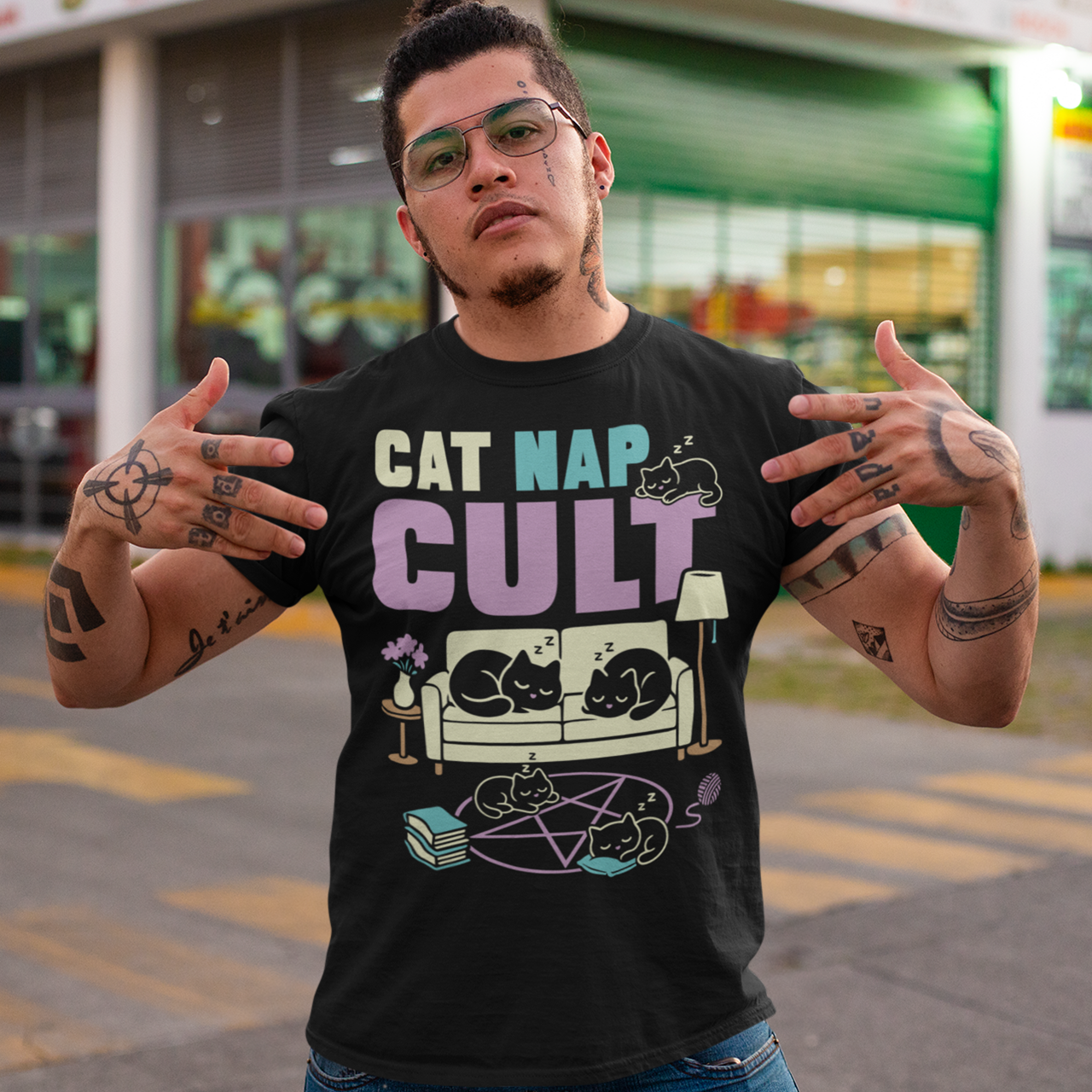 Black t-shirt with bold white text reading “Cat Nap Cult” above illustrations of sleeping cats lounging on a couch, books, and a pentagram-style ritual circle. The design includes cozy home details like a lamp, flowers, and yarn ball, creating a funny mix of occult humor and sleepy cat vibes in a minimalist monochrome style.