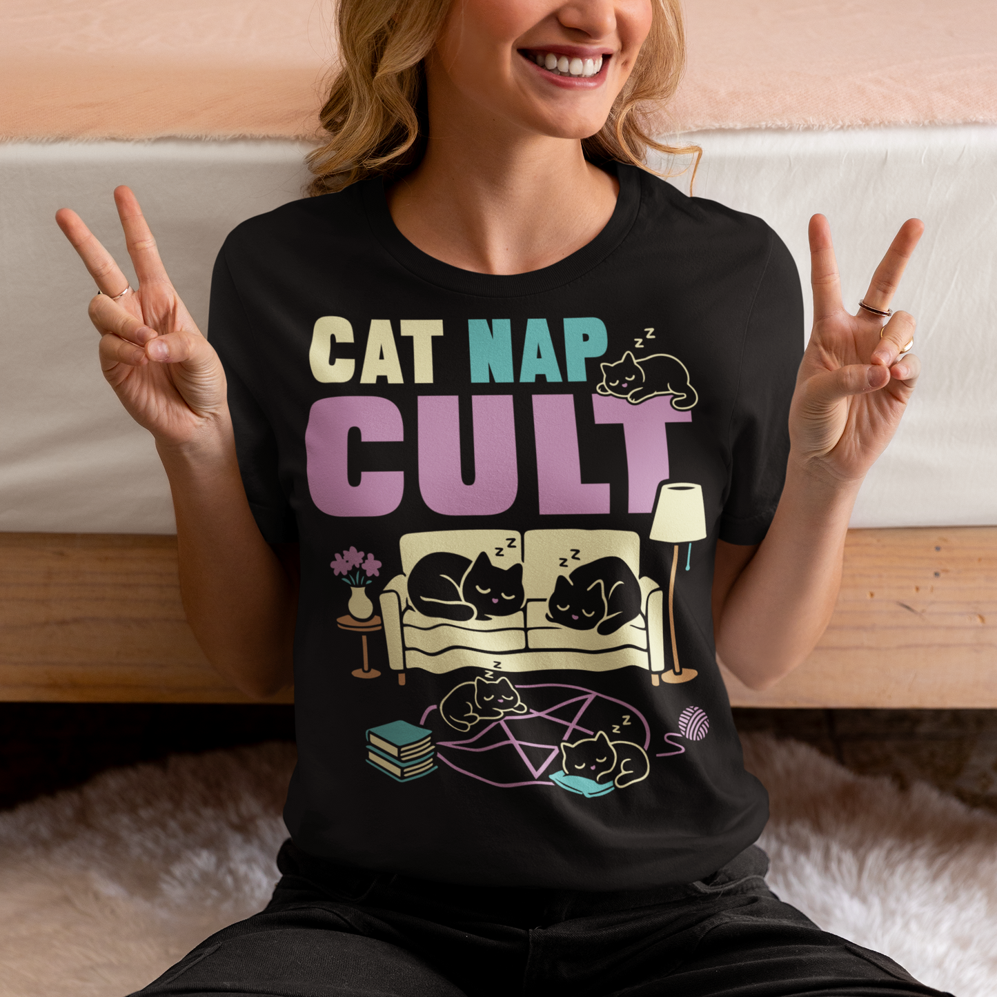 Black t-shirt with bold white text reading “Cat Nap Cult” above illustrations of sleeping cats lounging on a couch, books, and a pentagram-style ritual circle. The design includes cozy home details like a lamp, flowers, and yarn ball, creating a funny mix of occult humor and sleepy cat vibes in a minimalist monochrome style.