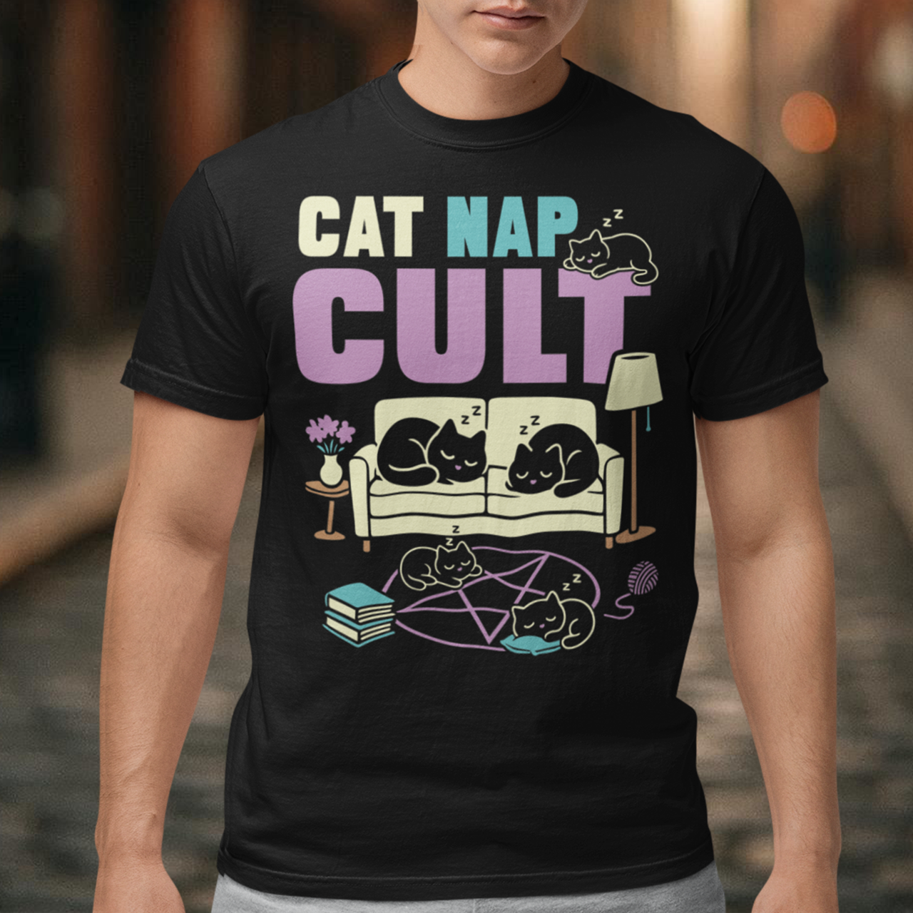 Black t-shirt with bold white text reading “Cat Nap Cult” above illustrations of sleeping cats lounging on a couch, books, and a pentagram-style ritual circle. The design includes cozy home details like a lamp, flowers, and yarn ball, creating a funny mix of occult humor and sleepy cat vibes in a minimalist monochrome style.