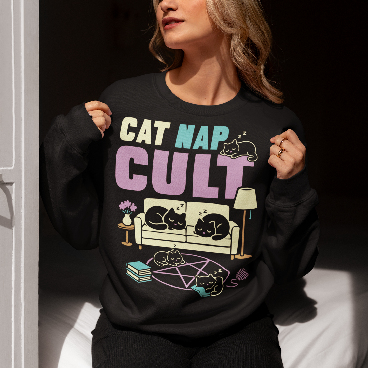 Black sweatshirt with bold white text reading “Cat Nap Cult” above illustrations of sleeping cats lounging on a couch, books, and a pentagram-style ritual circle. The design includes cozy home details like a lamp, flowers, and yarn ball, creating a funny mix of occult humor and sleepy cat vibes in a minimalist monochrome style.
