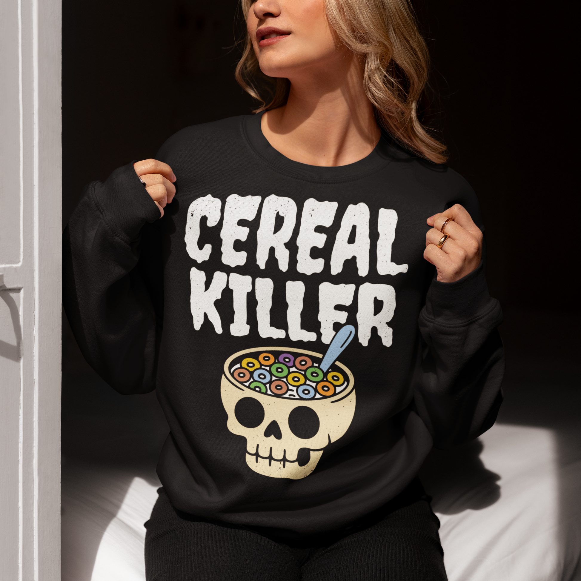 Black sweatshirt featuring the phrase “Cereal Killer” in large distressed white letters above a cartoon skull used as a cereal bowl filled with colorful cereal loops and a spoon. Funny dark humor design combining spooky Halloween style with breakfast pun humor.