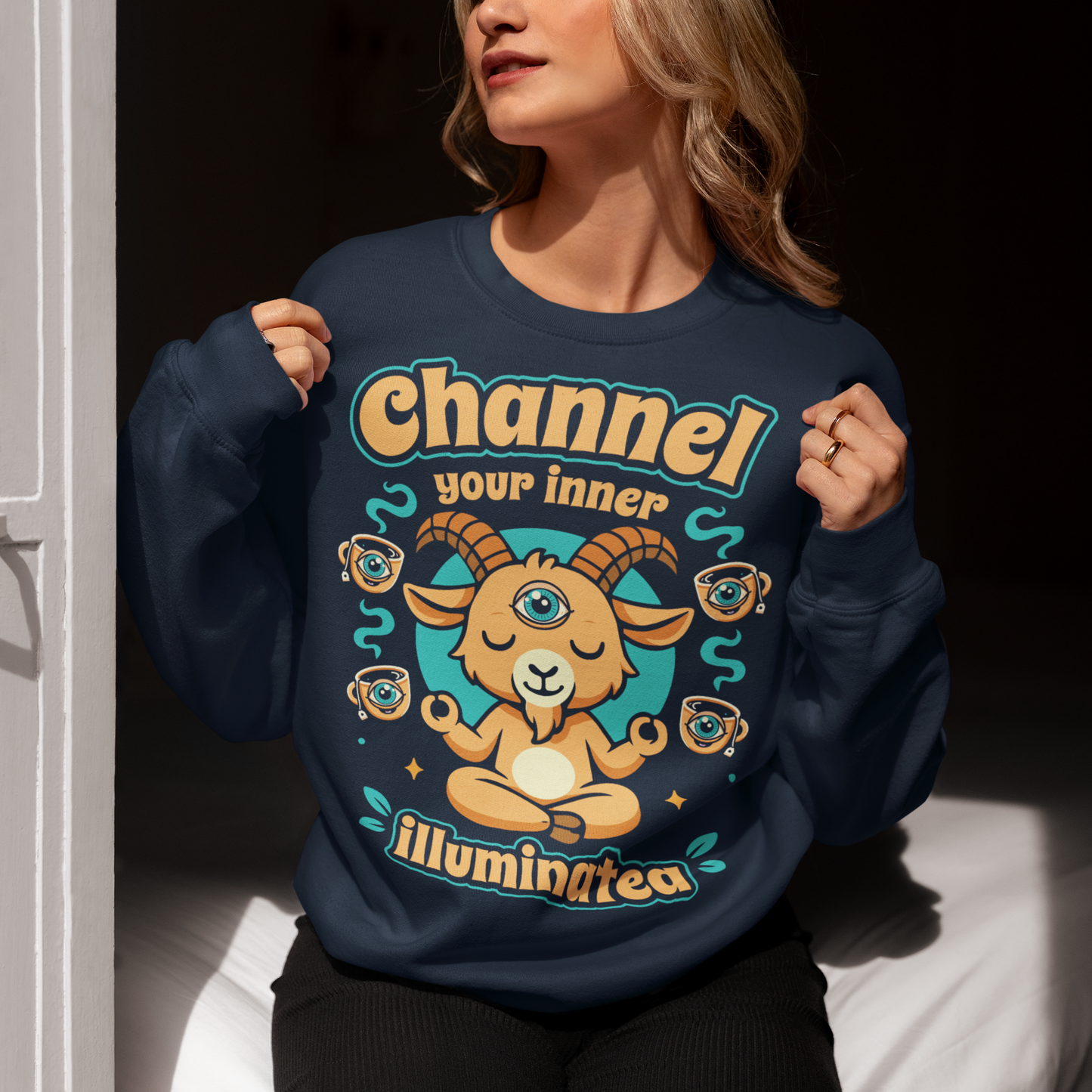 Navy t-shirt featuring a humorous occult-inspired design with the text “Channel Your Inner Illuminatea.” The artwork shows a meditating goat with a third eye, crossed legs, and raised hands, surrounded by floating teacups decorated with all-seeing eyes. Teal and tan accents create a mystical aesthetic blending spiritual symbolism, conspiracy humor, meditation themes, and playful occult artwork. Perfect for fans of witchy, esoteric, and funny graphic tees.