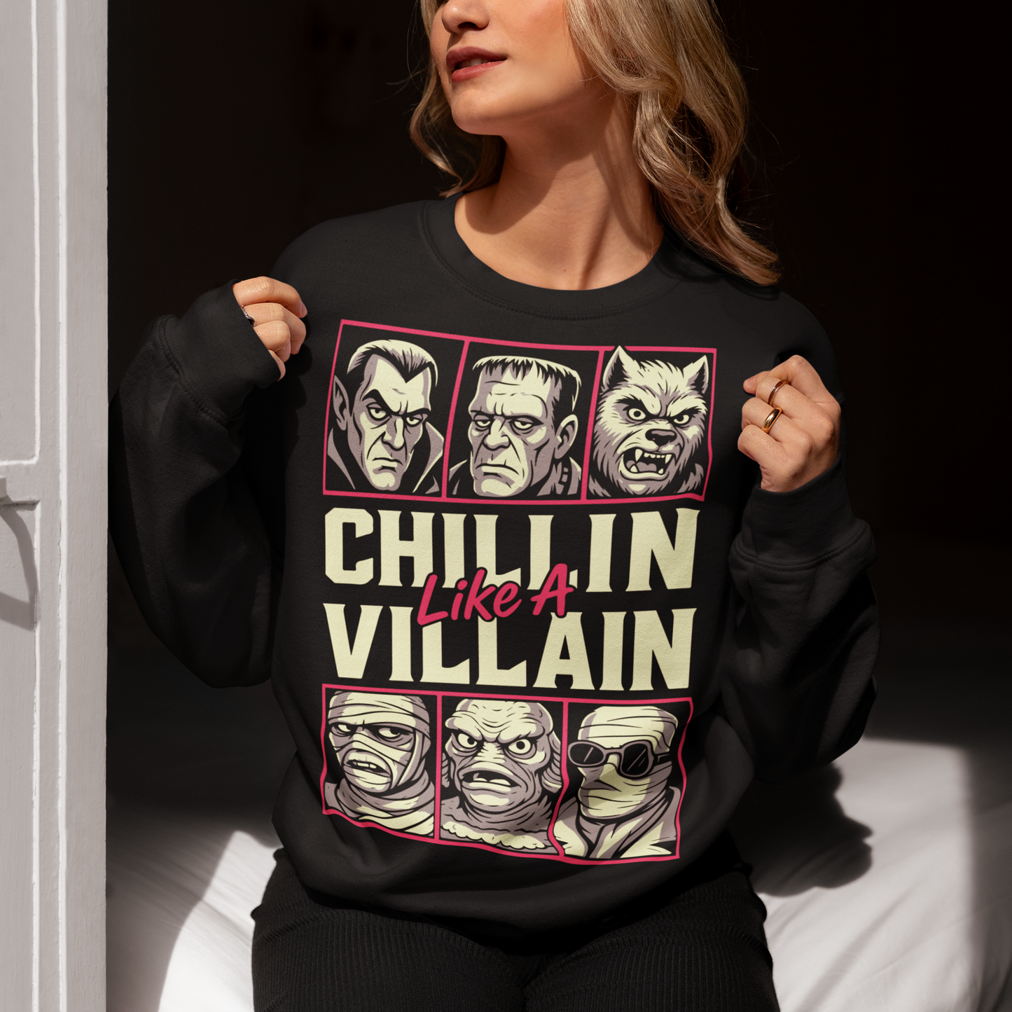 Black sweatshirt featuring the phrase "Chillin Like A Villain" with six classic horror monster portraits, including a vampire, Frankenstein's monster, werewolf, mummy, swamp creature, and the Invisible Man. The retro-inspired graphic uses distressed vintage styling with bold cream and pink lettering, making it a fun horror-themed shirt for Halloween, monster movie fans, goth fashion lovers, and classic horror enthusiasts.