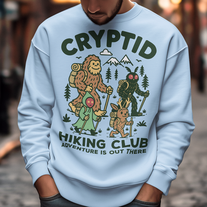 Light blue sweatshirt featuring a playful “Cryptid Hiking Club” design with Bigfoot, Mothman, flatwoods monster, and a jackalope hiking together through a forest trail. The artwork includes mountains, pine trees, backpacks, and trekking poles with the slogan “Adventure Is Out There.” A fun outdoor-themed graphic perfect for cryptid fans, hikers, campers, paranormal enthusiasts, and lovers of folklore-inspired apparel.