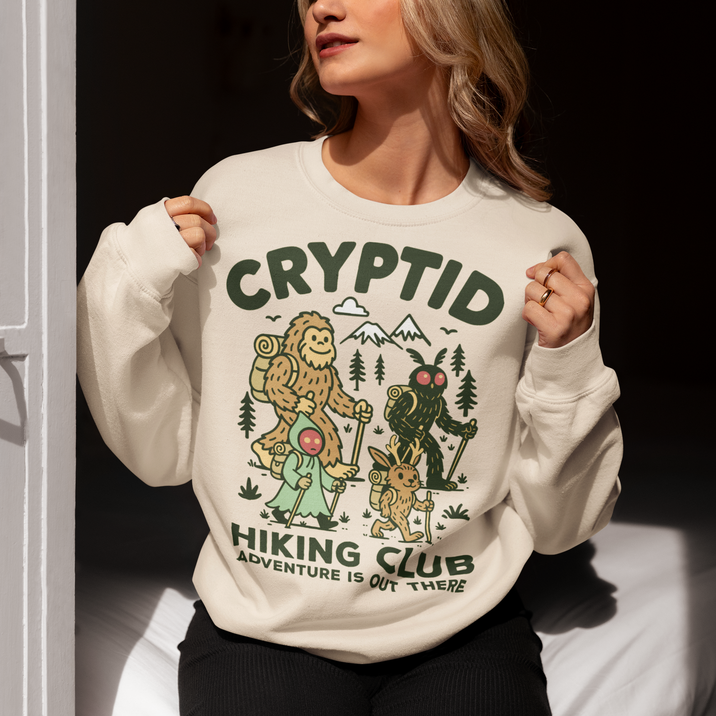 Beige sweatshirt featuring a playful “Cryptid Hiking Club” design with Bigfoot, Mothman, flatwoods monster, and a jackalope hiking together through a forest trail. The artwork includes mountains, pine trees, backpacks, and trekking poles with the slogan “Adventure Is Out There.” A fun outdoor-themed graphic perfect for cryptid fans, hikers, campers, paranormal enthusiasts, and lovers of folklore-inspired apparel.