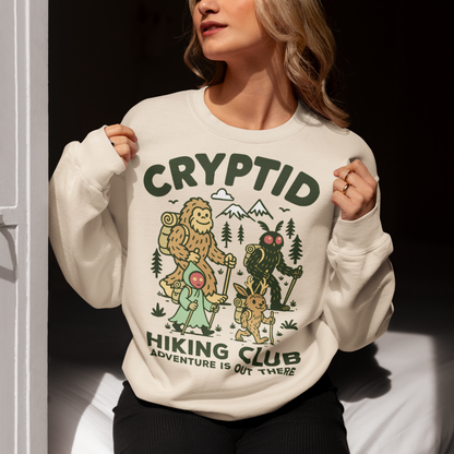 Beige sweatshirt featuring a playful “Cryptid Hiking Club” design with Bigfoot, Mothman, flatwoods monster, and a jackalope hiking together through a forest trail. The artwork includes mountains, pine trees, backpacks, and trekking poles with the slogan “Adventure Is Out There.” A fun outdoor-themed graphic perfect for cryptid fans, hikers, campers, paranormal enthusiasts, and lovers of folklore-inspired apparel.
