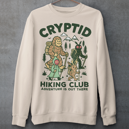 Beige sweatshirt featuring a playful “Cryptid Hiking Club” design with Bigfoot, Mothman, flatwoods monster, and a jackalope hiking together through a forest trail. The artwork includes mountains, pine trees, backpacks, and trekking poles with the slogan “Adventure Is Out There.” A fun outdoor-themed graphic perfect for cryptid fans, hikers, campers, paranormal enthusiasts, and lovers of folklore-inspired apparel.