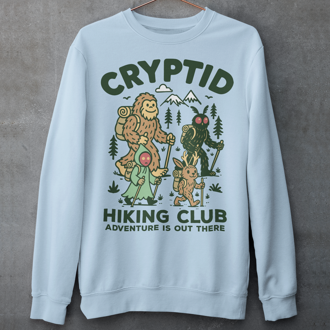 Light blue sweatshirt featuring a playful “Cryptid Hiking Club” design with Bigfoot, Mothman, flatwoods monster, and a jackalope hiking together through a forest trail. The artwork includes mountains, pine trees, backpacks, and trekking poles with the slogan “Adventure Is Out There.” A fun outdoor-themed graphic perfect for cryptid fans, hikers, campers, paranormal enthusiasts, and lovers of folklore-inspired apparel.