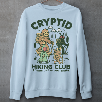 Light blue sweatshirt featuring a playful “Cryptid Hiking Club” design with Bigfoot, Mothman, flatwoods monster, and a jackalope hiking together through a forest trail. The artwork includes mountains, pine trees, backpacks, and trekking poles with the slogan “Adventure Is Out There.” A fun outdoor-themed graphic perfect for cryptid fans, hikers, campers, paranormal enthusiasts, and lovers of folklore-inspired apparel.
