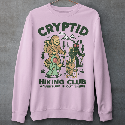 Pink sweatshirt featuring a playful “Cryptid Hiking Club” design with Bigfoot, Mothman, flatwoods monster, and a jackalope hiking together through a forest trail. The artwork includes mountains, pine trees, backpacks, and trekking poles with the slogan “Adventure Is Out There.” A fun outdoor-themed graphic perfect for cryptid fans, hikers, campers, paranormal enthusiasts, and lovers of folklore-inspired apparel.