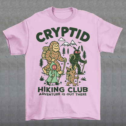 Pink t-shirt featuring a playful “Cryptid Hiking Club” design with Bigfoot, Mothman, flatwoods monster, and a jackalope hiking together through a forest trail. The artwork includes mountains, pine trees, backpacks, and trekking poles with the slogan “Adventure Is Out There.” A fun outdoor-themed graphic perfect for cryptid fans, hikers, campers, paranormal enthusiasts, and lovers of folklore-inspired apparel.