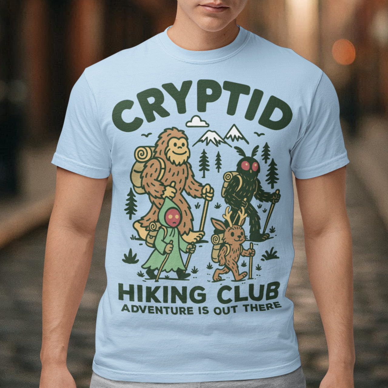 Light blue t-shirt featuring a playful “Cryptid Hiking Club” design with Bigfoot, Mothman, flatwoods monster, and a jackalope hiking together through a forest trail. The artwork includes mountains, pine trees, backpacks, and trekking poles with the slogan “Adventure Is Out There.” A fun outdoor-themed graphic perfect for cryptid fans, hikers, campers, paranormal enthusiasts, and lovers of folklore-inspired apparel.