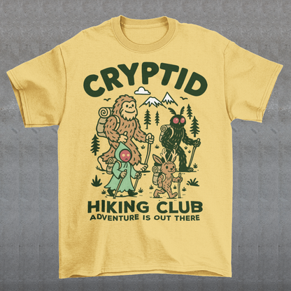 Yellow t-shirt featuring a playful “Cryptid Hiking Club” design with Bigfoot, Mothman, flatwoods monster, and a jackalope hiking together through a forest trail. The artwork includes mountains, pine trees, backpacks, and trekking poles with the slogan “Adventure Is Out There.” A fun outdoor-themed graphic perfect for cryptid fans, hikers, campers, paranormal enthusiasts, and lovers of folklore-inspired apparel.