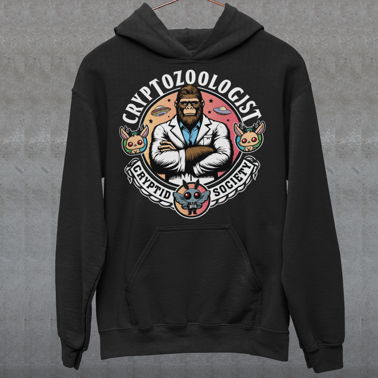 Black hoodie of a Bigfoot scientist with UFOs in the background. Mothman underneath and text above CRYPTOZOOLOGIST.