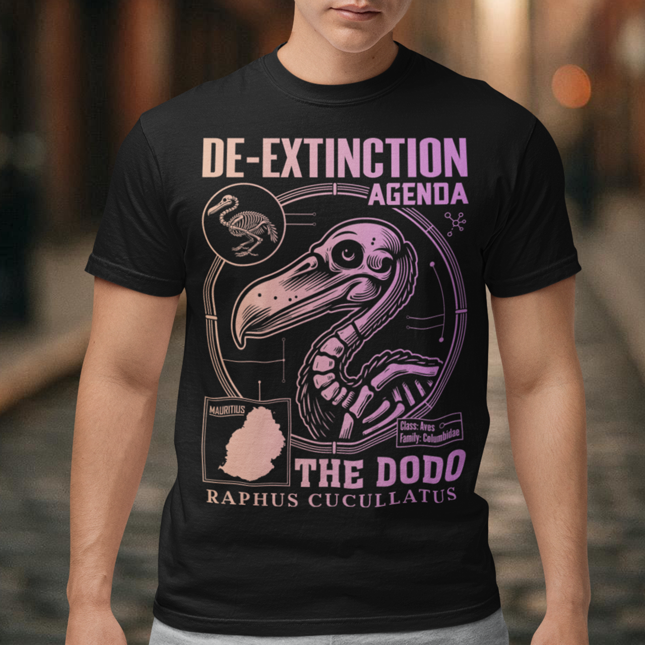 A graphic t-shirt featuring a stylized illustration of a dodo bird, standing upright with a proud, almost defiant expression. The dodo is depicted in vibrant colors, blending realism with a slightly cartoonish flair. Above the bird, bold, capitalized text reads "DE-EXTINCTION AGENDA" in a futuristic font, suggesting a science fiction or activist tone. The design evokes themes of genetic engineering, conservation, and the revival of extinct species. The shirt is a statement piece with humor.
