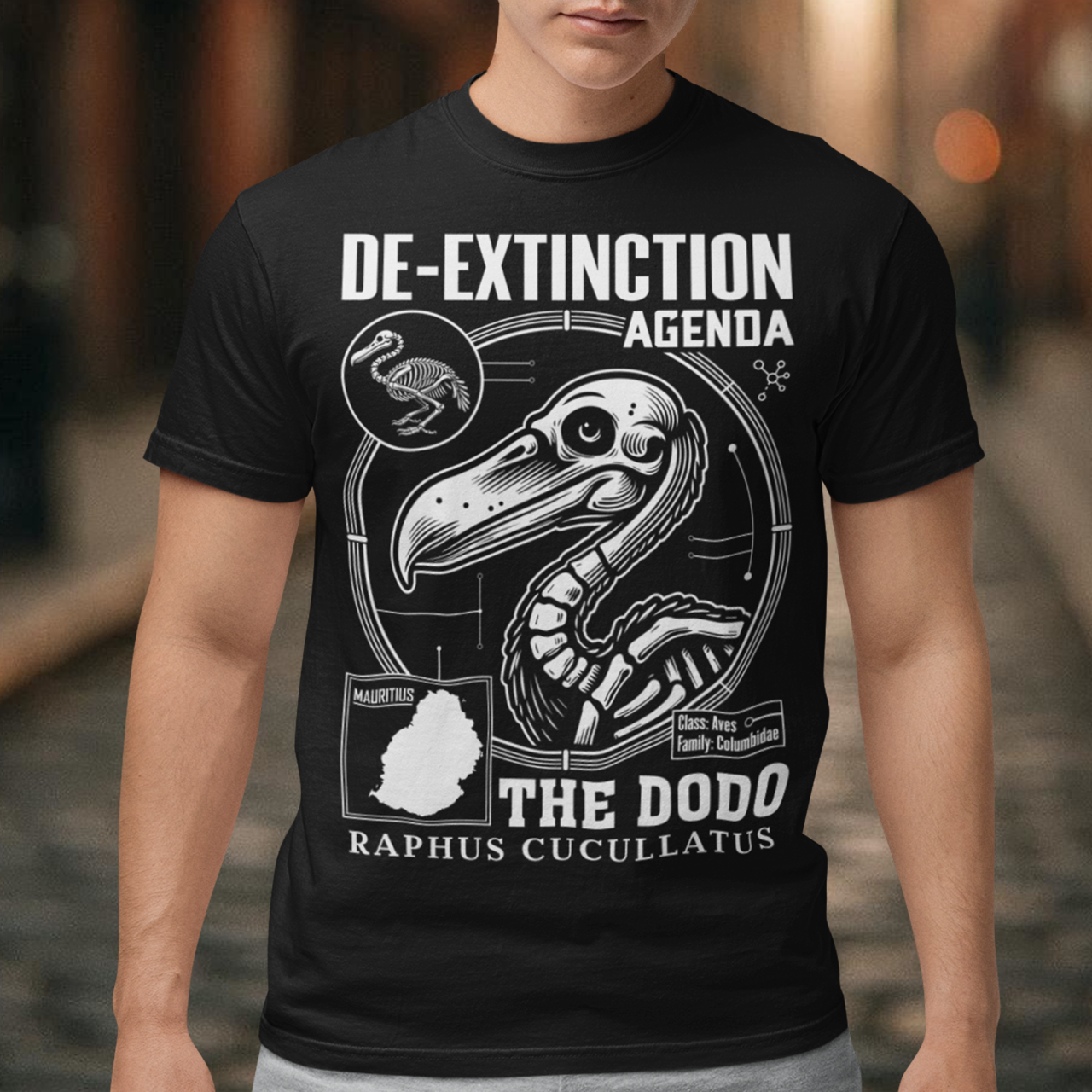 A graphic t-shirt featuring a stylized illustration of a dodo bird, standing upright with a proud, almost defiant expression. The dodo is depicted in vibrant colors, blending realism with a slightly cartoonish flair. Above the bird, bold, capitalized text reads "DE-EXTINCTION AGENDA" in a futuristic font, suggesting a science fiction or activist tone. The design evokes themes of genetic engineering, conservation, and the revival of extinct species. The shirt is a statement piece with humor.