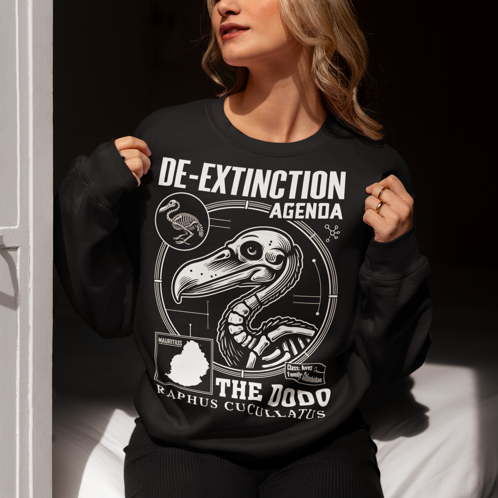 A graphic sweatshirt featuring a stylized illustration of a dodo bird, standing upright with a proud, almost defiant expression. The dodo is depicted in vibrant colors, blending realism with a slightly cartoonish flair. Above the bird, bold, capitalized text reads "DE-EXTINCTION AGENDA" in a futuristic font, suggesting a science fiction or activist tone. The design evokes themes of genetic engineering, conservation, and the revival of extinct species. The shirt is a statement piece with humor.