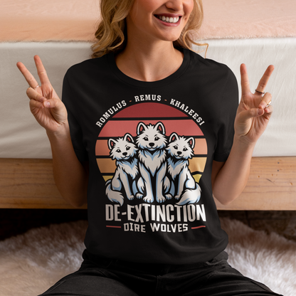 A graphic t-shirt design featuring three majestic Dire Wolves sitting side by side, facing forward with alert expressions. Behind them is a large, stylized retro sun composed of horizontal stripes in warm tones of orange, red, and yellow, evoking a vintage 80s aesthetic. Beneath the wolves, bold text reads “DE-EXTINCTION,” suggesting the return of these prehistoric predators. The design blends prehistoric imagery with nostalgic retro-futurism.