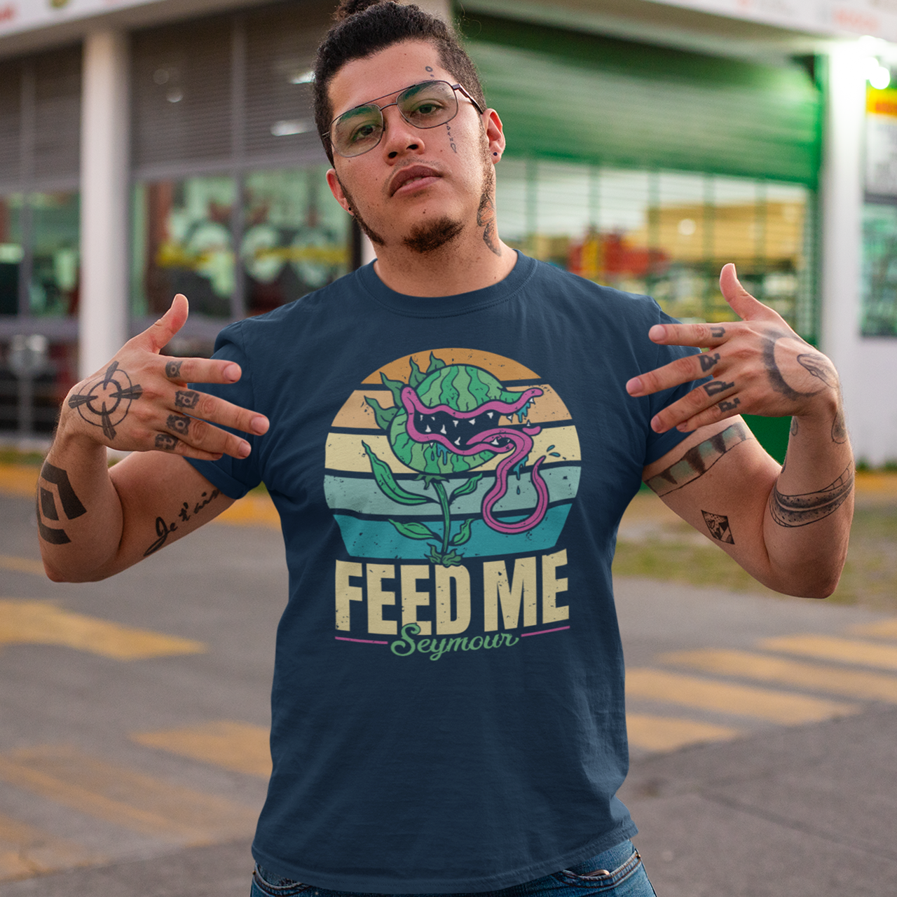 Navy t-shirt of sentient carnivorous plant with a retro sun background with text underneath "FEED ME SEYMOUR".