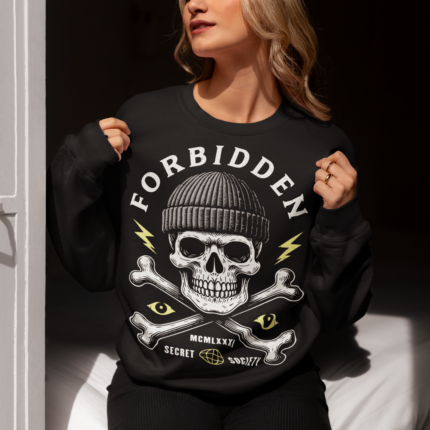 At the center sits a stark skull in a knit beanie, framed by crossed bones, watchful eyes, and crackling lightning bolts. Bold lettering reads Forbidden, with subtle details like Roman numerals and “Secret Society” giving the design the air of an underground emblem or outlaw insignia. The muted cream and acid-yellow accents pop against the black fabric, creating a gritty, modern occult aesthetic.