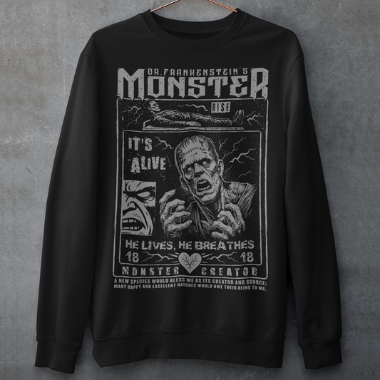 Black horror-themed sweatshirt featuring a vintage Frankenstein monster graphic in distressed gray print. The design includes comic-style panels with lightning bolts, Dr. Frankenstein’s monster rising from a lab table, and bold text reading “It’s Alive.” Gothic typography and retro horror artwork give the shirt a classic monster movie aesthetic, perfect for Halloween, horror fans, gothic fashion lovers, and collectors of vintage spooky apparel.