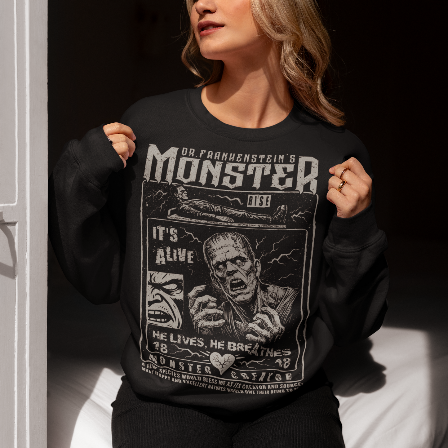 Black horror-themed sweatshirt featuring a vintage Frankenstein monster graphic in distressed gray print. The design includes comic-style panels with lightning bolts, Dr. Frankenstein’s monster rising from a lab table, and bold text reading “It’s Alive.” Gothic typography and retro horror artwork give the shirt a classic monster movie aesthetic, perfect for Halloween, horror fans, gothic fashion lovers, and collectors of vintage spooky apparel.