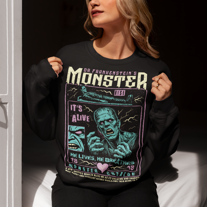 Black horror-themed sweatshirt featuring a vintage Frankenstein monster graphic in distressed gray print. The design includes comic-style panels with lightning bolts, Dr. Frankenstein’s monster rising from a lab table, and bold text reading “It’s Alive.” Gothic typography and retro horror artwork give the shirt a classic monster movie aesthetic, perfect for Halloween, horror fans, gothic fashion lovers, and collectors of vintage spooky apparel.