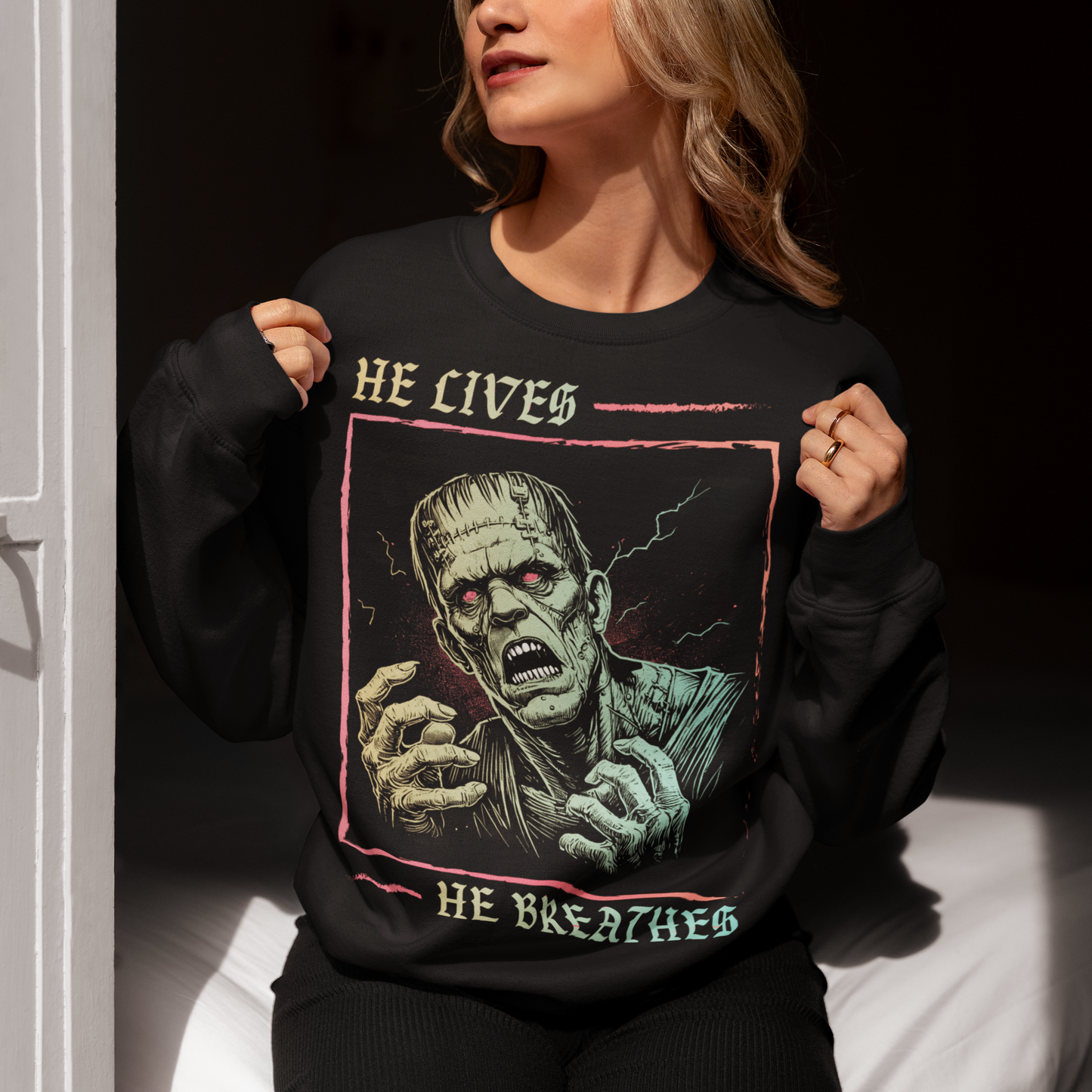 Black sweatshirt of Frankenstein with text above HE LIVES and text below HE BREATHES.