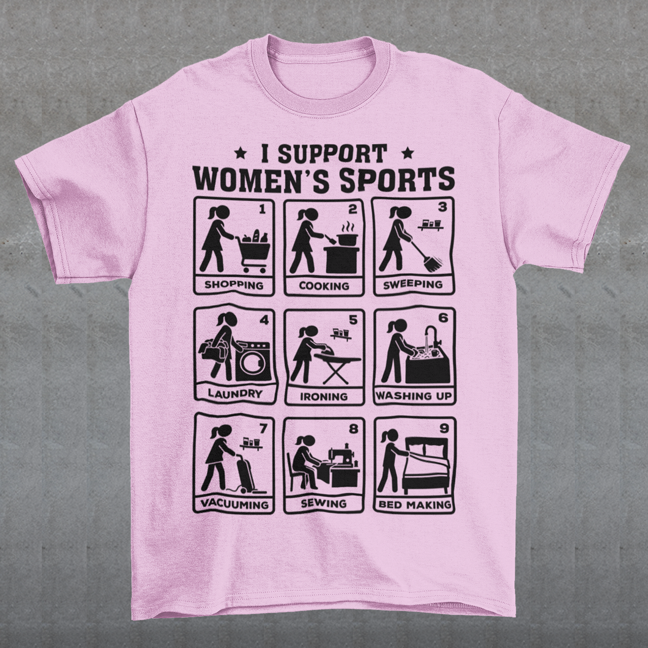 Pink t-shirt featuring the humorous phrase “I SUPPORT WOMEN’S SPORTS” with nine illustrated panels parodying different household activities: shopping, cooking, sweeping, laundry, ironing, washing up, vacuuming, sewing, and bed making. Black graphics create a retro sports-style design with a sarcastic, tongue-in-cheek theme.