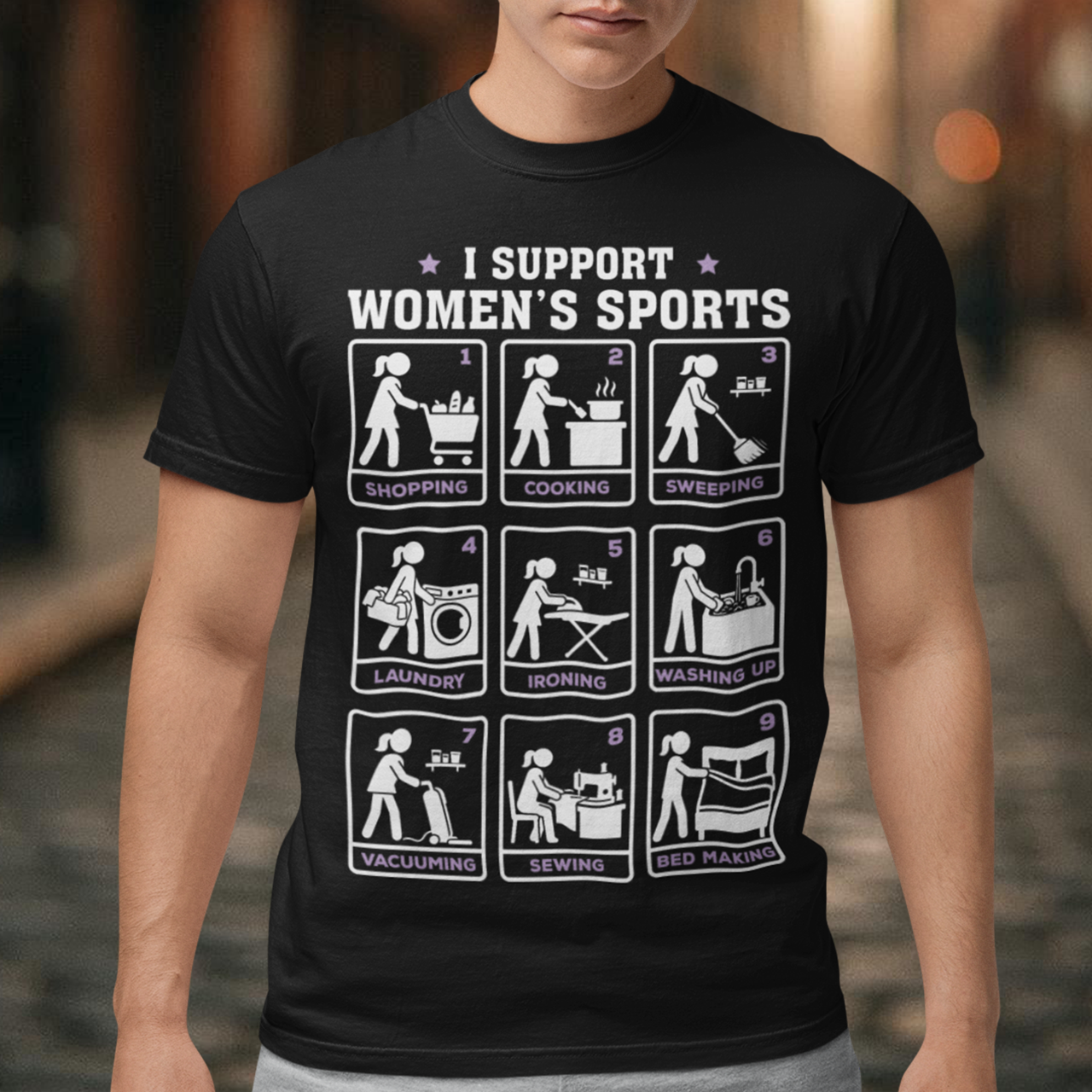 Black t-shirt featuring the humorous phrase “I SUPPORT WOMEN’S SPORTS” with nine illustrated panels parodying different household activities: shopping, cooking, sweeping, laundry, ironing, washing up, vacuuming, sewing, and bed making. White and purple graphics create a retro sports-style design with a sarcastic, tongue-in-cheek theme.