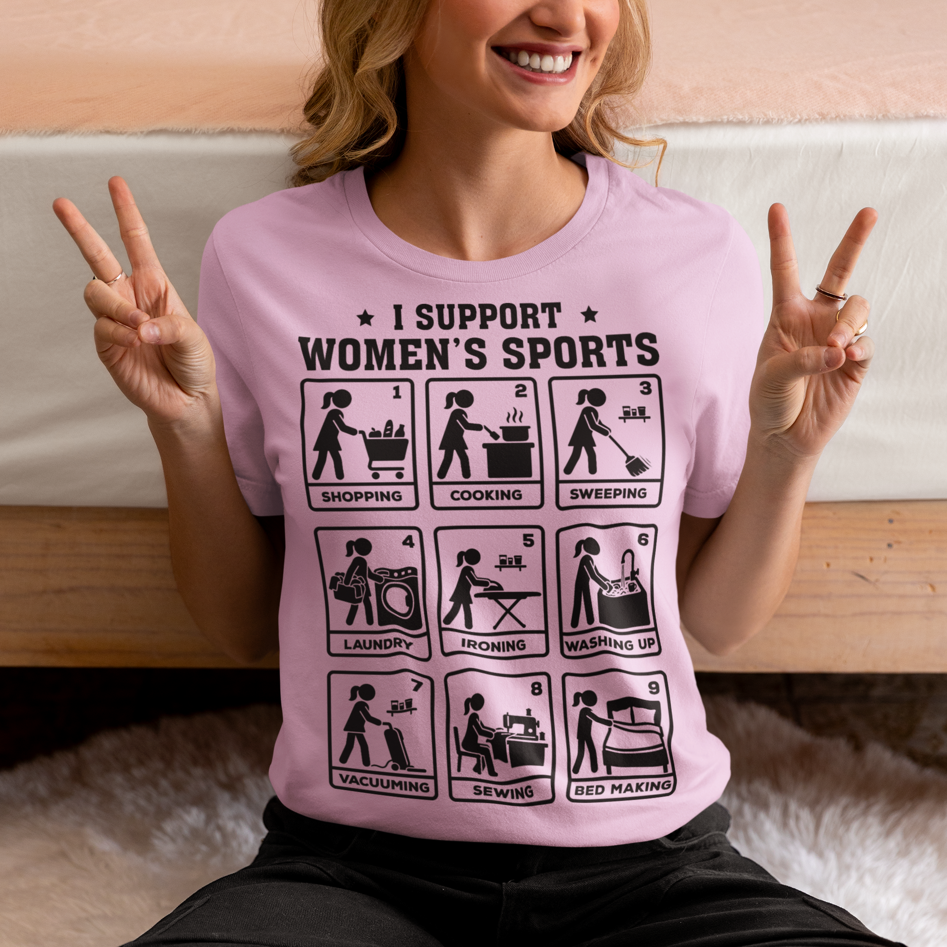 Pink t-shirt featuring the humorous phrase “I SUPPORT WOMEN’S SPORTS” with nine illustrated panels parodying different household activities: shopping, cooking, sweeping, laundry, ironing, washing up, vacuuming, sewing, and bed making. Black graphics create a retro sports-style design with a sarcastic, tongue-in-cheek theme.