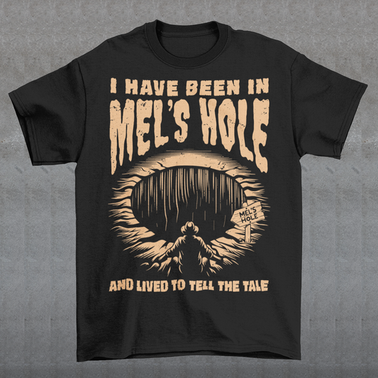 Black t-shirt featuring a vintage horror-style design with the text “I Have Been In Mel’s Hole And Lived To Tell The Tale.” The graphic shows a shadowy figure standing before a deep mysterious pit labeled “Mel’s Hole.” Distressed tan lettering and eerie artwork give the shirt a cryptid, paranormal, and conspiracy-inspired aesthetic perfect for fans of strange legends, spooky humor, and occult mystery designs.