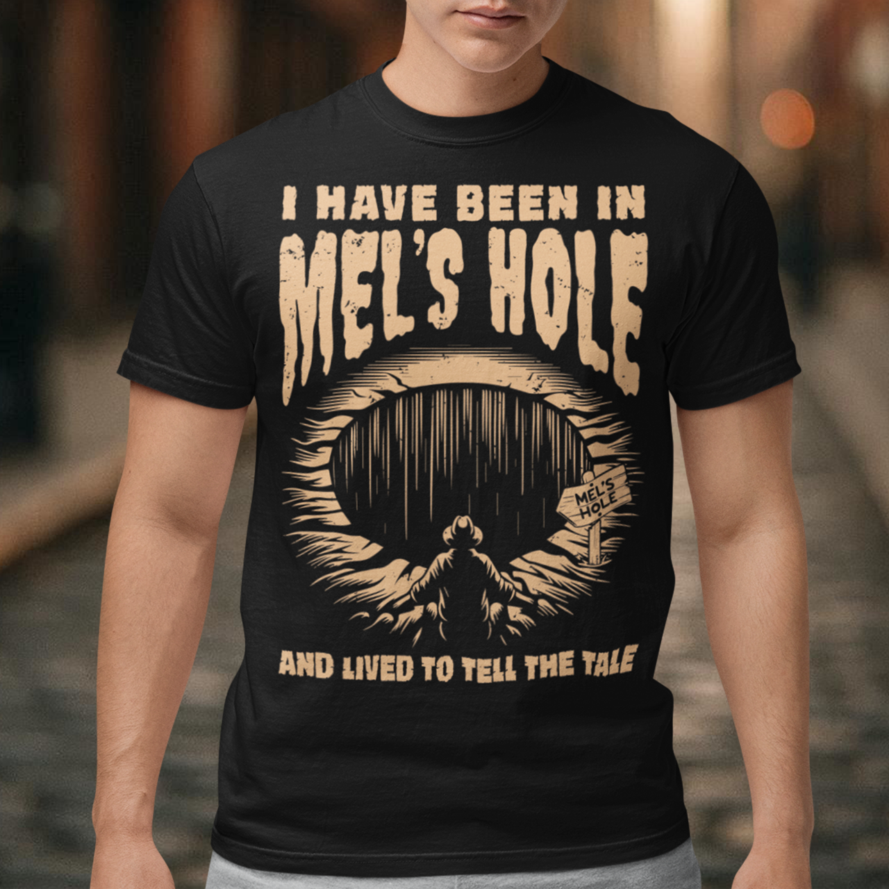 Black t-shirt featuring a vintage horror-style design with the text “I Have Been In Mel’s Hole And Lived To Tell The Tale.” The graphic shows a shadowy figure standing before a deep mysterious pit labeled “Mel’s Hole.” Distressed tan lettering and eerie artwork give the shirt a cryptid, paranormal, and conspiracy-inspired aesthetic perfect for fans of strange legends, spooky humor, and occult mystery designs.